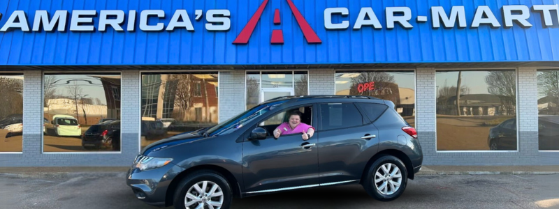 Car-Mart of Oxford General Manager, Pat Stutsy-Waldrep, giving a thumbs-up from the open window of a grey crossover in front of a dealership.