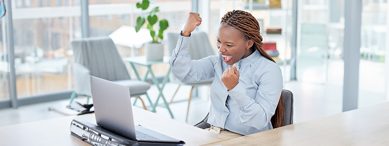 Woman sitting at desk looking at laptop with hands raised in the air